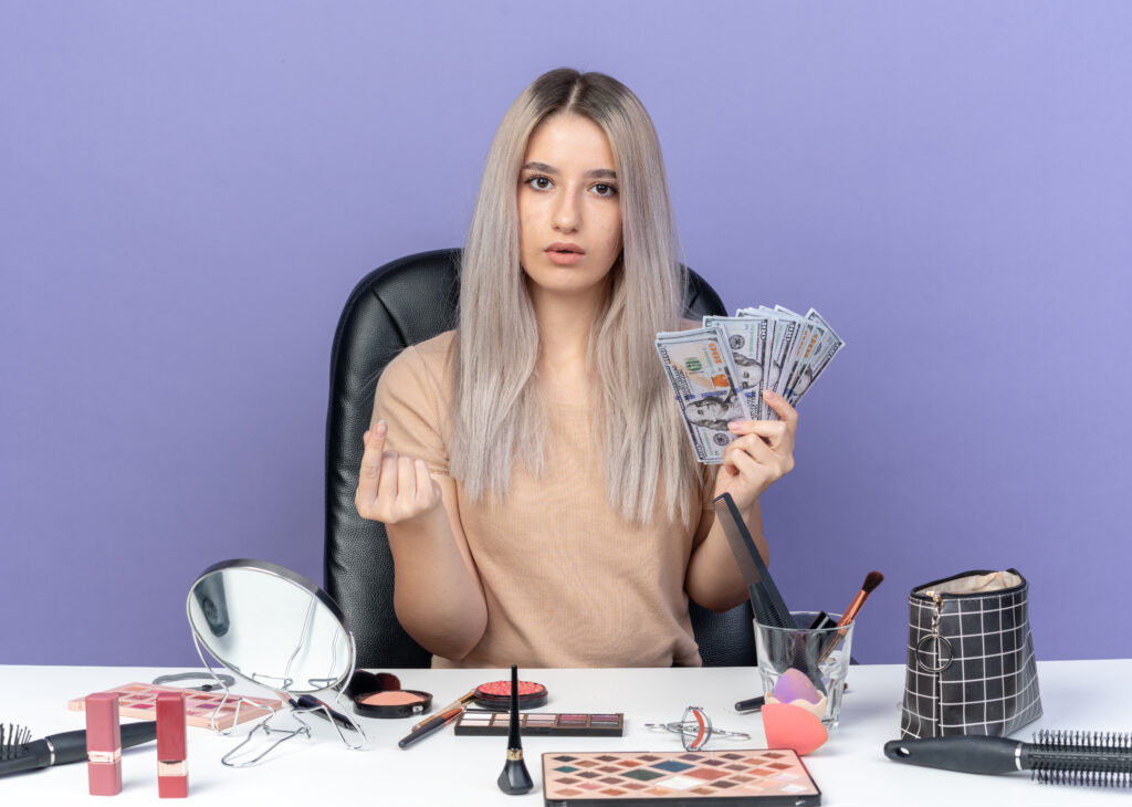Woman holding cash while sitting at a table with makeup tools, suggesting pay-for-play modeling or beauty industry costs